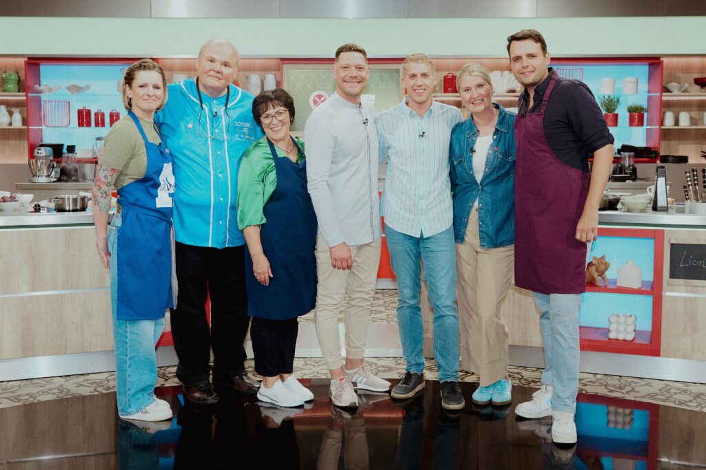 Ein Foto der Kandidaten und Kandidatinnen der ZDF Küchenschlacht. Im Vordergrund Robin Pietsch als Moderator, rechts und links von ihm jeweils drei Kandidaten der Woche. Im Hintergrund zu erkennen ist das Fernsehstudio der Küchenschlacht. 