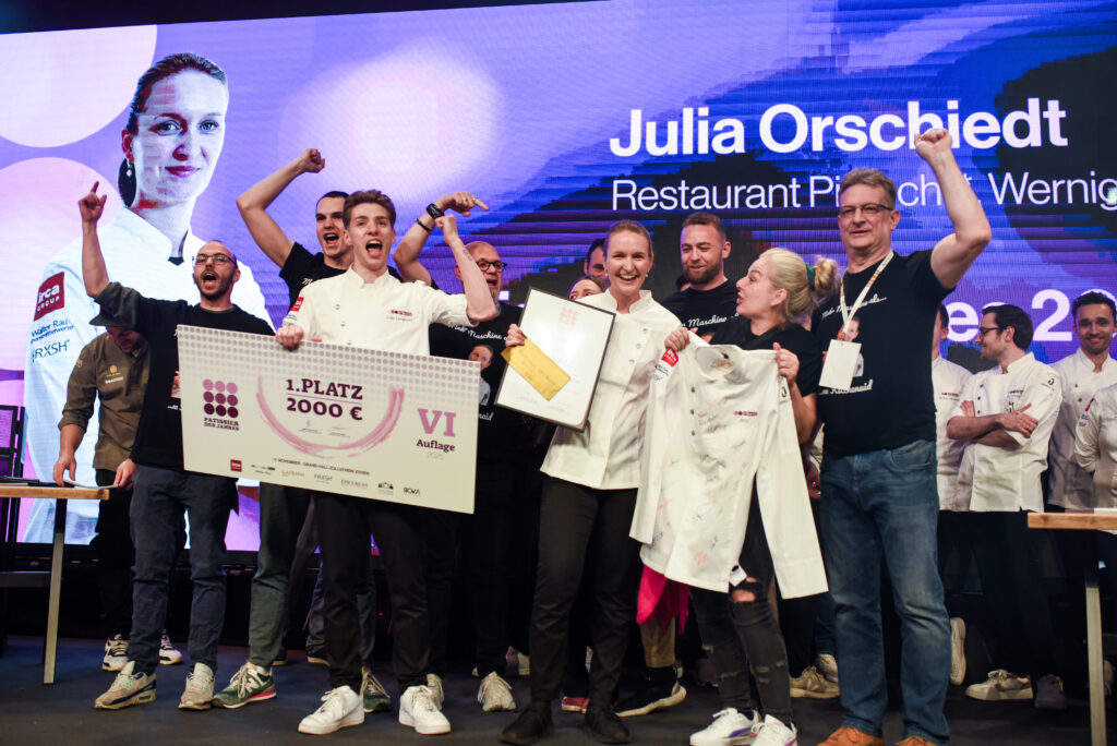 Julia Orschiedt, Gewinnerin des Wettbewerbs "Patissier des Jahres 2025" auf der Bühne, umgeben von ihrem Assistenten Luis Hendricks, dem Team des Restaurant pietsch in Wernigerode, Freunden und Familie